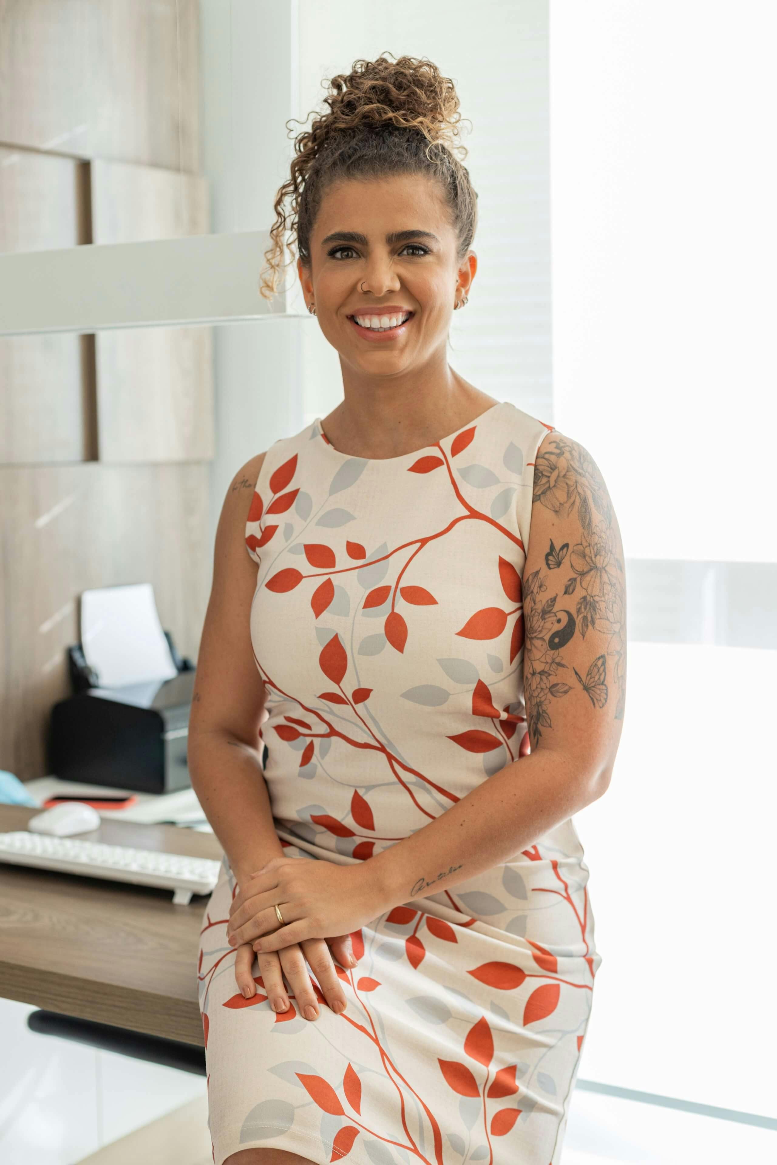 Foto profissional da Dra. Naiara Galvão de pé em seu consultório, com um vestido florido branco com detalhes vermelhos, exibindo um largo e caloroso sorriso.